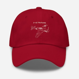 May include: A red baseball cap featuring a white outline of a P-40 Warhawk airplane. The text "P-40 Warhawk" is above the plane graphic. The cap is made of a soft material and has a curved brim.