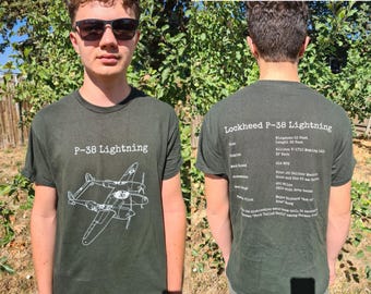 May include: Dark green t-shirt featuring a white illustration of a P-38 Lightning aircraft. The front of the shirt displays the text "P-38 Lightning." The back of the shirt has the text "Lockheed P-38 Lightning" and aircraft specifications.