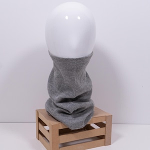 Może przedstawiać: Szary dzianinowy ocieplacz na szyję, prezentowany na białej głowie manekina. Ocieplacz na szyję wykonany jest z miękkiego, teksturowanego materiału i jest narzucony na małą drewnianą skrzynkę. Ocieplacz na szyję jest w jednolitym szarym kolorze.