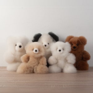 May include: Five plush teddy bears in various colors, including white, brown, black, and beige. The bears are sitting on a wooden surface.