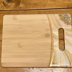 May include: A rectangular bamboo cutting board with a white and gold epoxy resin inlay. The inlay features a swirl pattern and a cutout handle. The resin is accented with small, clear crystals.
