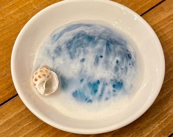 May include: A white ceramic dish with a blue and white swirl design. A small seashell is resting on the edge of the dish.