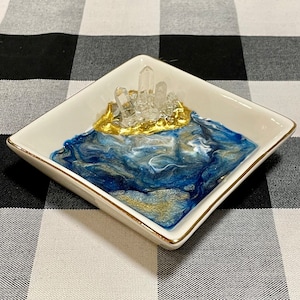 May include: A white ceramic trinket dish with a gold rim and a blue and white resin design. The dish features a cluster of clear quartz crystals on top of a gold accent.