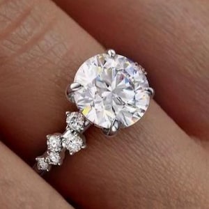 Może przedstawiać: Srebrny pierścionek zaręczynowy z dużym, okrągłym, czystym diamentem centralnym i mniejszymi diamentami osadzonymi w oprawie typu pavé.