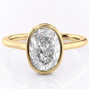 Może przedstawiać: Złoty pierścionek z diamentem o owalnym szlifie. Diament osadzony jest w oprawie typu bezel, a obrączka jest z gładkiego złota. Pierścionek jest prezentowany na białym tle, podkreślając jego blask i design. Design pierścionka jest prosty i elegancki.