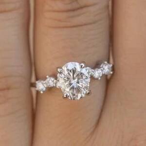 Może przedstawiać: Srebrny pierścionek zaręczynowy z dużą, okrągłą, przezroczystą szlachetnym kamieniem jako centralnym elementem. Mniejsze, przezroczyste kamienie szlachetne są osadzone wzdłuż obrączki, dodając blasku. Pierścionek jest na palcu.