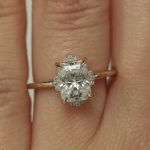 Może przedstawiać: Złoty pierścionek z dużym, prostokątnym, przezroczystym kamieniem szlachetnym, otoczonym mniejszymi kamieniami. Pierścionek ma delikatną złotą obrączkę, tworząc klasyczny i elegancki wygląd. Ten pierścionek jest symbolem zaangażowania i miłości.