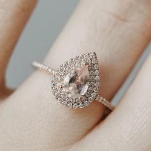 May include: A rose gold engagement ring with a pear-shaped, pale peach gemstone, encircled by a halo of small, clear stones. The ring sits on a slender band, presenting a classic and elegant design.