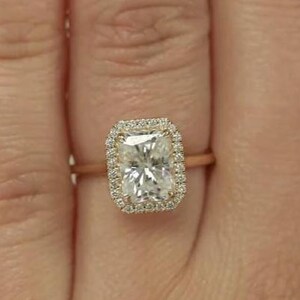 Może przedstawiać: Pierścionek z różowego złota z prostokątnym, przezroczystym kamieniem centralnym otoczonym halo z mniejszych kamieni. Pierścionek osadzony jest na delikatnej obrączce, podkreślającej błyszczący design. To biżuteria.