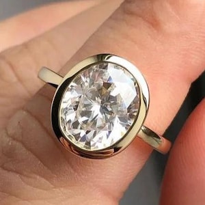 Może przedstawiać: Złoty pierścionek z dużym diamentem o owalnym kształcie. Diament osadzony jest w oprawie typu bezel, a obrączka jest z gładkiego złota. Pierścionek jest prezentowany na palcu, podkreślając jego rozmiar i blask.