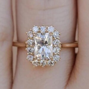 Może przedstawiać: Złoty pierścionek z prostokątnym, przezroczystym kamieniem szlachetnym otoczonym mniejszymi, okrągłymi kamieniami szlachetnymi. Pierścionek osadzony jest na cienkiej, złotej obrączce, prezentując klasyczny wzór halo. Pierścionek to biżuteria.