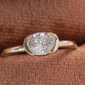 Może przedstawiać: Złoty pierścionek z dużym, owalnym diamentem w oprawie. Pierścionek jest prezentowany w brązowym, aksamitnym pudełku. Biżuteria, która podkreśla blask diamentu.