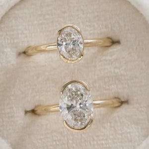 Könnte beinhalten: Zwei goldene Ringe mit oval geschliffenen Diamanten werden in einer weißen Samt-Ringbox präsentiert. Die Diamanten sind klar und funkelnd, gefasst in einem zarten Goldband. Die Ringe sind mittig in der Box platziert.