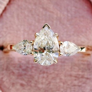 Może przedstawiać: Pierścionek zaręczynowy z diamentem w kształcie gruszki, z dwoma mniejszymi diamentowymi gruszkami po obu stronach, osadzony w złotej obrączce. Pierścionek jest prezentowany w różowym aksamitnym pudełku na pierścionki.