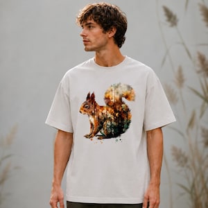 May include: A light grey short-sleeve t-shirt featuring a watercolour-style graphic of a squirrel. The squirrel illustration incorporates warm orange, brown, and dark green tones, with a forest-like pattern integrated into its fur and bushy tail.