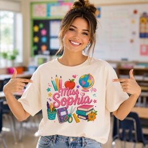 May include: A cream-coloured t-shirt featuring a colourful graphic print of school supplies including pencils, an apple, a globe, books, a notebook, scissors, and a ruler, with the text "Miss Sophia" in large pink script lettering.