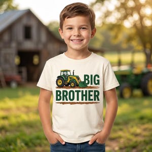 May include: A cream-coloured short-sleeve t-shirt featuring a graphic of a green and yellow tractor with a front loader. The text "BIG BROTHER" is printed in bold green capital letters below the tractor, with a brown graphic of dirt underneath.