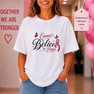 May include: A white t-shirt featuring the black and pink text Emma's Believe in Hope, accented with two small butterflies, a pink awareness ribbon, and tiny hearts. A pink tag hangs to the right with the text Customize it, add your name.
