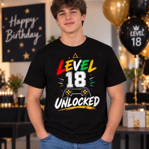 May include: A black short-sleeved t-shirt featuring a graphic print with the text "LEVEL 18 UNLOCKED" in white, red, green, and yellow. The design includes a stylised video game controller icon and colourful accents on the front of the garment.