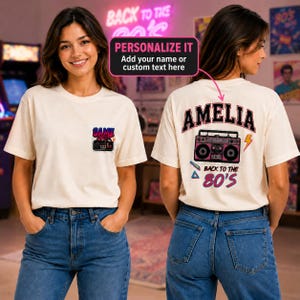 May include: A cream-coloured t-shirt featuring a small graphic on the front left chest with a game controller and the text "GAME OVER." The back displays a large boombox graphic, the name "AMELIA," and the text "BACK TO THE 80'S" with geometric shapes.
