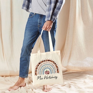 May include: A beige tote bag with a rainbow design featuring a red apple and the words "Teach Lead Inspire Support Encourage Listen Care" in a brown leopard print. The text "Miss Wellsburg" is printed in black below the rainbow.
