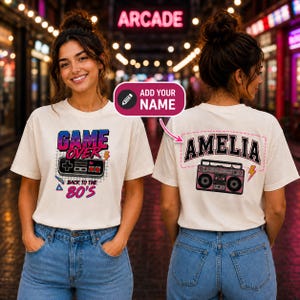 May include: A cream-coloured t-shirt featuring a front graphic with a video game controller and the text "GAME OVER BACK TO THE 80'S" and a back graphic with a boombox, a lightning bolt, and the customisable name "AMELIA" in a varsity font.