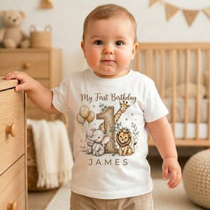 May include: A white short-sleeved t-shirt featuring a graphic of a giraffe, an elephant, and a lion with balloons and the text My First Birthday JAMES. The design includes a large number one and green foliage accents on the front of the shirt.