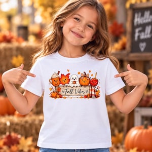 May include: A white t-shirt featuring a graphic illustration of a smiling ghost, a pumpkin, a mug of hot cocoa, a maple leaf, and a candy apple resting on a wooden sign with the text Fall Vibes written in a cursive black font.