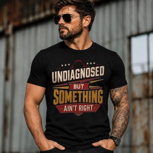 May include: A black short-sleeved t-shirt featuring a graphic print with the text "UNDIAGNOSED BUT SOMETHING AIN'T RIGHT" in distressed white and yellow lettering, accented by red banners and small white stars on a dark background.