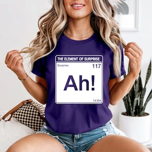 May include: A dark purple t-shirt with the text "THE ELEMENT OF SURPRISE" and the word "Ah!" in a white box. The shirt also features the word "Surprise" and the number "117".