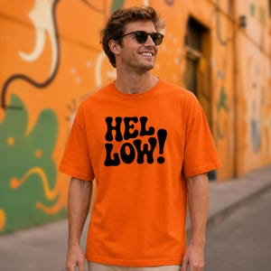 May include: A bright orange short-sleeved t-shirt featuring the text "HEL LOW!" printed in a bold, black, wavy, retro-style font across the chest. The shirt has a crew neckline and a standard fit, providing a simple and clean appearance.