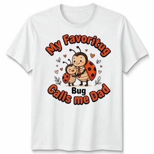 May include: White t-shirt with a cartoon ladybug illustration. The design shows a large and small ladybug hugging, with the text "My Favourite Bug Calls me Dad" in orange and black. Small hearts and leaves surround the ladybugs.