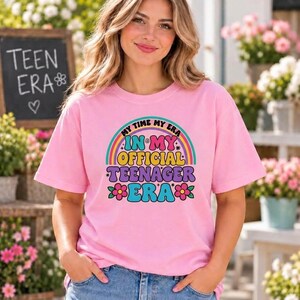 May include: A light pink short-sleeve t-shirt featuring a colourful graphic print of a rainbow and flowers with the text "MY TIME MY ERA IN MY OFFICIAL TEENAGER ERA". The design uses bold yellow, pink, and blue lettering with star accents.