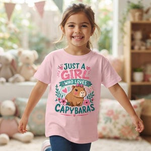 May include: A light pink short-sleeved t-shirt featuring a graphic of a brown capybara with a pink bow. The text reads JUST A GIRL WHO LOVES CAPYBARAS, surrounded by pink hearts, green leaves, and pink flowers in a decorative, colourful design.