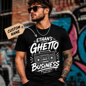May include: A black short-sleeve t-shirt featuring a white graphic print of a boombox with the text "ETHAN'S GHETTO BUSINESS" and "BUILT DIFFERENT - STAYS LOUD" below it. A tan rectangular tag with the text "CUSTOM NAME" is attached to the shirt.