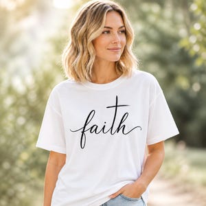 May include: A white short-sleeved t-shirt featuring the word faith printed in black cursive script across the chest, where the letter t is stylised as a cross. The garment has a crew neckline and a relaxed fit, made of a solid white fabric.