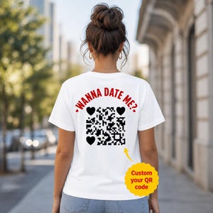 May include: A white short-sleeved t-shirt featuring the red text "WANNA DATE ME?" arched above a black QR code surrounded by three black hearts. A yellow circular graphic with the text "Custom your QR code" points to the QR code.