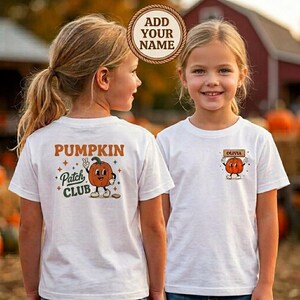 May include: A white t-shirt featuring a graphic on the back with the text PUMPKIN PATCH CLUB and a cartoon pumpkin character. The front of the shirt displays a smaller graphic of a pumpkin character holding a sign with the name OLIVIA.