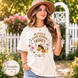 May include: A cream-coloured t-shirt featuring a graphic print with the text 'OLIVIA'S KIND WORDS ARE LIKE HONEY' and 'Proverbs 16:24'. The design includes a honeybee, a honeycomb, and colourful floral illustrations surrounding the central text.
