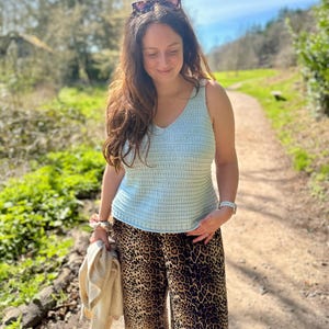 May include: A light blue crochet tank top paired with leopard print pants. The outfit is completed with a cream-colored sweater and sunglasses. The person is walking on a path in a park.