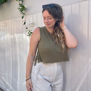 May include: A woman wearing a green sleeveless knitted crop top with white pants. The top has a tie detail on the sides.