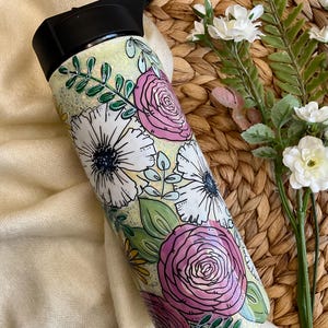 Puede incluir: Una botella de agua con estampado floral y tapa negra. La botella presenta un fondo verde claro con flores blancas y rosas y hojas verdes. El diseño está dibujado a mano y la botella es cilíndrica.