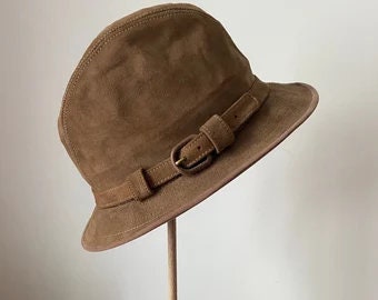 May include: A brown suede cloche hat with a buckled band. The hat has a rounded crown and a slightly angled brim. The band is secured with a rectangular buckle. The hat is displayed on a wooden stand.