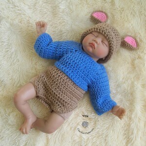 May include: A crocheted baby outfit in blue and brown with a matching crocheted hat featuring pink ears. The hat has a brown crocheted brim and pink crocheted ears. The outfit has a blue crocheted top and a brown crocheted bottom. The outfit is laid out on a white fluffy surface.