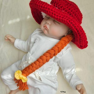 May include: A red crochet hat with a wide brim and a long orange braid, adorned with a yellow bow. The hat is worn by a baby dressed in a white onesie. The image is taken from above.