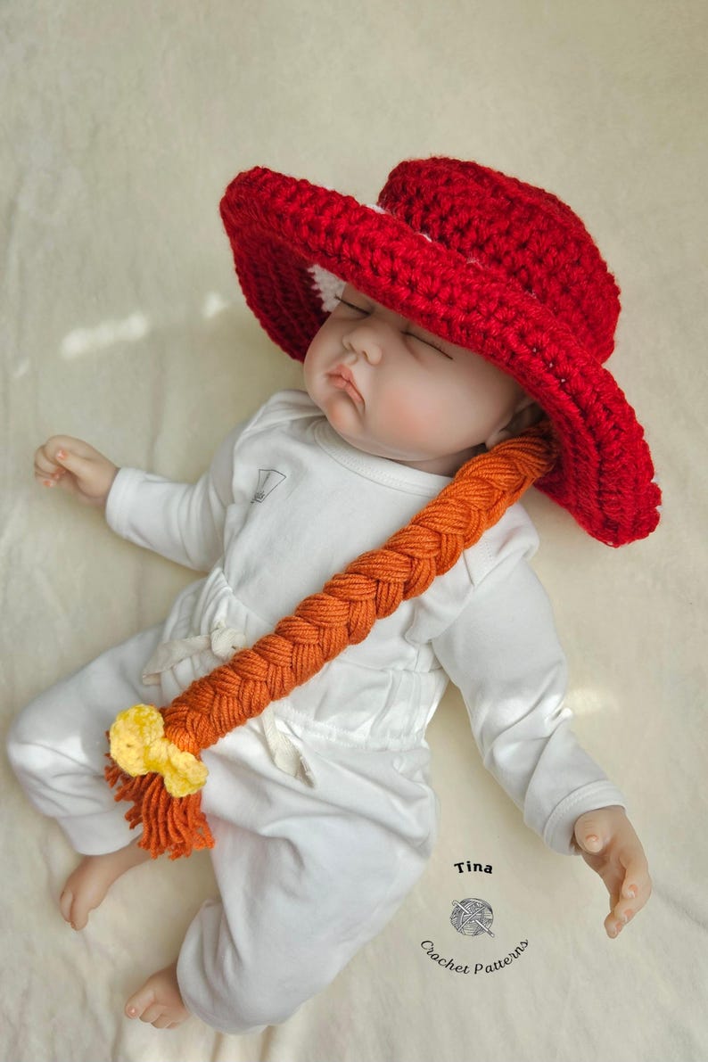 May include: A red crochet hat with a wide brim and a long orange braid, adorned with a yellow bow. The hat is worn by a baby dressed in a white onesie. The image is taken from above.
