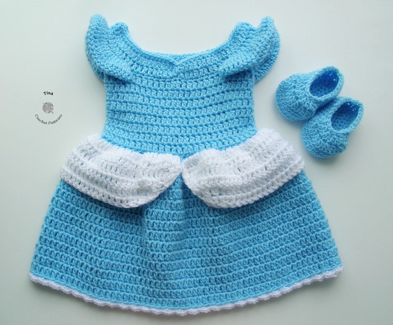 Peut inclure: Une robe de b&eacute;b&eacute; en crochet bleu clair avec des bordures blanches et des chaussures en crochet assorties. La robe a une jupe &agrave; volants et un d&eacute;collet&eacute; en c&oelig;ur.