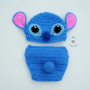 May include: A blue crocheted baby hat and diaper set with pink ears, blue eyes, and a black nose. The set is designed to look like Stitch from the Disney movie Lilo & Stitch.