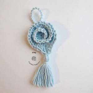 May include: A light blue crocheted flower with a tassel hanging from the bottom. The flower is made of multiple layers of crocheted yarn and has a small loop at the top for hanging.