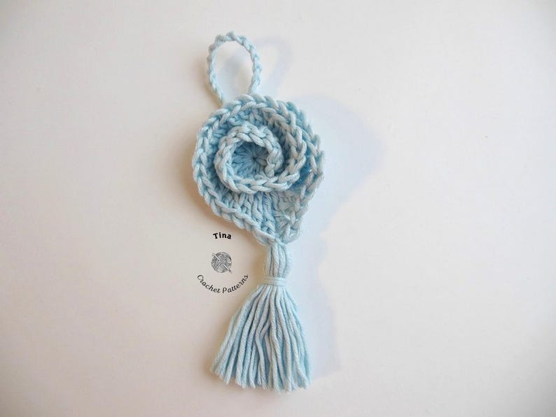 May include: A light blue crocheted flower with a tassel hanging from the bottom. The flower is made of multiple layers of crocheted yarn and has a small loop at the top for hanging.